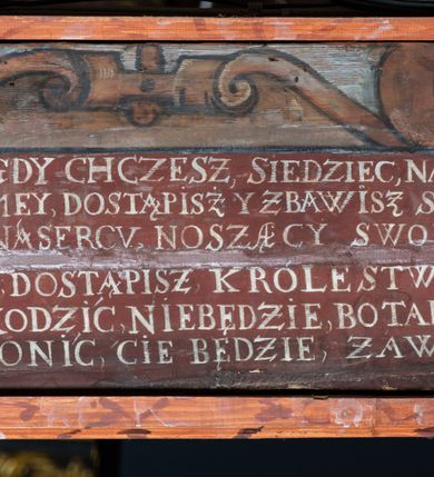 Zdjęcie nr 11: Rzeźba pełna, Chrystus przybity do krzyża trzema gwoździami w lekkim zwisie z głową silnie skręconą w prawo. Twarz podłużna z otwartymi ustami i półprzymkniętymi oczami, okolona kędzierzawą, ciemną brodą; włosy długie spływające na plecy i prawe ramię. Nogi delikatnie ugięte w kolanach; stopy przebite jednym gwoździem, w układzie prawa na lewą. Na głowie Chrystus ma założoną zieloną koronę cierniową. Perizonium w formie tkaniny przewiązanej diagonalnie przez biodra, ze zwisem na prawym boku. Sylwetka wychudzona, smukła, o delikatnym skręcie bioder, z wyraźnie zaznaczonym użyleniem i czerwono-sinymi śladami męki. Polichromia naturalistyczna w partiach ciała, o bladej kolorystyce bieli i błękitu oraz intensywnej czerwonej krwi; perizonium złocone. Nad głową Chrystusa zawieszony zielono-czarny titulus w formie kartusza z napisem „INRI”, ozdobiony motywem małżowinowo-chrząstkowym. Krzyż drewniany, prosty, malowany na czarno. U dołu krzyża zawieszone cztery wota w kształcie złotych serduszek przypiętych czerwonymi kokardkami.