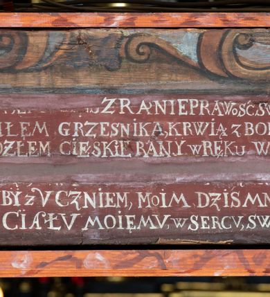 Zdjęcie nr 12: Rzeźba pełna, Chrystus przybity do krzyża trzema gwoździami w lekkim zwisie z głową silnie skręconą w prawo. Twarz podłużna z otwartymi ustami i półprzymkniętymi oczami, okolona kędzierzawą, ciemną brodą; włosy długie spływające na plecy i prawe ramię. Nogi delikatnie ugięte w kolanach; stopy przebite jednym gwoździem, w układzie prawa na lewą. Na głowie Chrystus ma założoną zieloną koronę cierniową. Perizonium w formie tkaniny przewiązanej diagonalnie przez biodra, ze zwisem na prawym boku. Sylwetka wychudzona, smukła, o delikatnym skręcie bioder, z wyraźnie zaznaczonym użyleniem i czerwono-sinymi śladami męki. Polichromia naturalistyczna w partiach ciała, o bladej kolorystyce bieli i błękitu oraz intensywnej czerwonej krwi; perizonium złocone. Nad głową Chrystusa zawieszony zielono-czarny titulus w formie kartusza z napisem „INRI”, ozdobiony motywem małżowinowo-chrząstkowym. Krzyż drewniany, prosty, malowany na czarno. U dołu krzyża zawieszone cztery wota w kształcie złotych serduszek przypiętych czerwonymi kokardkami.