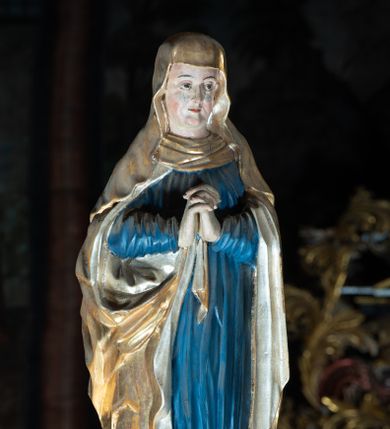Zdjęcie nr 1: Rzeźba pełna, Matka Boska w pozie stojącej na półkolistym cokole, ukazana frontalnie ze splecionymi na wysokości piersi dłońmi. Twarz pociągła, o delikatnie rzeźbionych rysach, dużych oczach ze wzrokiem skierowanym przed siebie, wyraźnie podkreślonym podbródku. Ubrana jest w niebieską, długą suknię drapowaną w równolegle, rurkowate fałdy i złoty płaszcz przerzucony przez ramiona ze srebrzonym podbiciem. Na głowie ma złoty welon opadający na ramiona. Polichromia w odsłoniętych partiach ciała naturalistyczna.