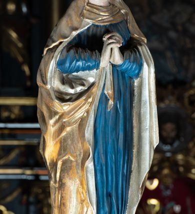 Zdjęcie nr 2: Rzeźba pełna, Matka Boska w pozie stojącej na półkolistym cokole, ukazana frontalnie ze splecionymi na wysokości piersi dłońmi. Twarz pociągła, o delikatnie rzeźbionych rysach, dużych oczach ze wzrokiem skierowanym przed siebie, wyraźnie podkreślonym podbródku. Ubrana jest w niebieską, długą suknię drapowaną w równolegle, rurkowate fałdy i złoty płaszcz przerzucony przez ramiona ze srebrzonym podbiciem. Na głowie ma złoty welon opadający na ramiona. Polichromia w odsłoniętych partiach ciała naturalistyczna.