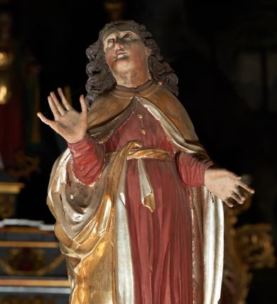 Zdjęcie nr 1: Rzeźba pełna, św. Jan ukazany w pozycji stojącej, frontalnie na półkolistym cokole, z głową zwróconą delikatnie w prawą stronę i uniesioną w górę; ręce ugięte w łokciach, prawa uniesiona. Twarz pełna, oczy zwrócone ku górze, włosy długie, brązowe, w postaci gęstych oddzielnych loków. Święty ubrany jest w długą, ciemnoczerwoną suknię, przepasaną w talii sznurem oraz złoty płaszcz ze srebrzoną podszewką, zapięty u szyi, opadający na ramiona i przewieszony prawym końcem przez pasek w talii. Polichromia w odsłoniętych partiach ciała naturalistyczna.