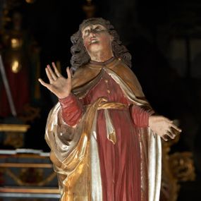 Zdjęcie nr 1: Rzeźba pełna, św. Jan ukazany w pozycji stojącej, frontalnie na półkolistym cokole, z głową zwróconą delikatnie w prawą stronę i uniesioną w górę; ręce ugięte w łokciach, prawa uniesiona. Twarz pełna, oczy zwrócone ku górze, włosy długie, brązowe, w postaci gęstych oddzielnych loków. Święty ubrany jest w długą, ciemnoczerwoną suknię, przepasaną w talii sznurem oraz złoty płaszcz ze srebrzoną podszewką, zapięty u szyi, opadający na ramiona i przewieszony prawym końcem przez pasek w talii. Polichromia w odsłoniętych partiach ciała naturalistyczna.