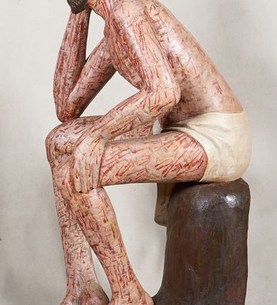 Zdjęcie nr 2: Figura pełnoplastyczna przedstawiająca Chrystusa Frasobliwego. Chrystus ukazany w pozycji siedzącej na drewnianym postumencie, wspierający prawą ręką głowę, a lewą opierający na lewym udzie. Ciało o silnie podkreślonej klatce piersiowej z wąską talią, bardzo szczupłe. Twarz podłużna z długim i wąskim nosem, z głęboko osadzonymi oczami, wzrokiem skierowanym w dół, okolona gęstą, krótką brodą, zawiniętą na końcu w dwa małe pukle. Figura wyrzeźbiona bez włosów. Na głowę założona peruka z długich, naturalnych, jasnych włosów, a na nich korona z przeplecionych gałązek. Perizonium rzeźbione, białe, ciasno oplatające biodra, z jednym końcem rozwianym pod prawym udem. Polichromia w odsłoniętych partiach ciała naturalistyczna. Spod korony cierniowej oraz po całym ciele spływają bardzo liczne, drobne strużki krwi oraz zaznaczone poziomymi kreskami sińce. Na rzeźbę nałożony jest współczesny czerwony płaszcz z materiału, spięty pod szyją metalową klamrą, na brzegach obszyty złotą bordiurą. 


