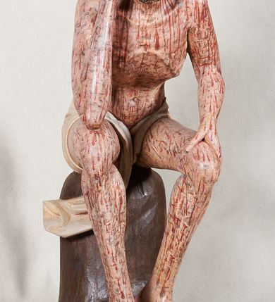 Zdjęcie nr 4: Figura pełnoplastyczna przedstawiająca Chrystusa Frasobliwego. Chrystus ukazany w pozycji siedzącej na drewnianym postumencie, wspierający prawą ręką głowę, a lewą opierający na lewym udzie. Ciało o silnie podkreślonej klatce piersiowej z wąską talią, bardzo szczupłe. Twarz podłużna z długim i wąskim nosem, z głęboko osadzonymi oczami, wzrokiem skierowanym w dół, okolona gęstą, krótką brodą, zawiniętą na końcu w dwa małe pukle. Figura wyrzeźbiona bez włosów. Na głowę założona peruka z długich, naturalnych, jasnych włosów, a na nich korona z przeplecionych gałązek. Perizonium rzeźbione, białe, ciasno oplatające biodra, z jednym końcem rozwianym pod prawym udem. Polichromia w odsłoniętych partiach ciała naturalistyczna. Spod korony cierniowej oraz po całym ciele spływają bardzo liczne, drobne strużki krwi oraz zaznaczone poziomymi kreskami sińce. Na rzeźbę nałożony jest współczesny czerwony płaszcz z materiału, spięty pod szyją metalową klamrą, na brzegach obszyty złotą bordiurą. 


