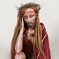 Zdjęcie nr 1: Figura pełnoplastyczna przedstawiająca Chrystusa Frasobliwego. Chrystus ukazany w pozycji siedzącej na drewnianym postumencie, wspierający prawą ręką głowę, a lewą opierający na lewym udzie. Ciało o silnie podkreślonej klatce piersiowej z wąską talią, bardzo szczupłe. Twarz podłużna z długim i wąskim nosem, z głęboko osadzonymi oczami, wzrokiem skierowanym w dół, okolona gęstą, krótką brodą, zawiniętą na końcu w dwa małe pukle. Figura wyrzeźbiona bez włosów. Na głowę założona peruka z długich, naturalnych, jasnych włosów, a na nich korona z przeplecionych gałązek. Perizonium rzeźbione, białe, ciasno oplatające biodra, z jednym końcem rozwianym pod prawym udem. Polichromia w odsłoniętych partiach ciała naturalistyczna. Spod korony cierniowej oraz po całym ciele spływają bardzo liczne, drobne strużki krwi oraz zaznaczone poziomymi kreskami sińce. Na rzeźbę nałożony jest współczesny czerwony płaszcz z materiału, spięty pod szyją metalową klamrą, na brzegach obszyty złotą bordiurą. 


