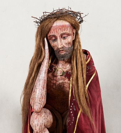 Zdjęcie nr 1: Figura pełnoplastyczna przedstawiająca Chrystusa Frasobliwego. Chrystus ukazany w pozycji siedzącej na drewnianym postumencie, wspierający prawą ręką głowę, a lewą opierający na lewym udzie. Ciało o silnie podkreślonej klatce piersiowej z wąską talią, bardzo szczupłe. Twarz podłużna z długim i wąskim nosem, z głęboko osadzonymi oczami, wzrokiem skierowanym w dół, okolona gęstą, krótką brodą, zawiniętą na końcu w dwa małe pukle. Figura wyrzeźbiona bez włosów. Na głowę założona peruka z długich, naturalnych, jasnych włosów, a na nich korona z przeplecionych gałązek. Perizonium rzeźbione, białe, ciasno oplatające biodra, z jednym końcem rozwianym pod prawym udem. Polichromia w odsłoniętych partiach ciała naturalistyczna. Spod korony cierniowej oraz po całym ciele spływają bardzo liczne, drobne strużki krwi oraz zaznaczone poziomymi kreskami sińce. Na rzeźbę nałożony jest współczesny czerwony płaszcz z materiału, spięty pod szyją metalową klamrą, na brzegach obszyty złotą bordiurą. 


