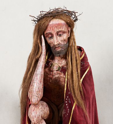 Zdjęcie nr 1: Figura pełnoplastyczna przedstawiająca Chrystusa Frasobliwego. Chrystus ukazany w pozycji siedzącej na drewnianym postumencie, wspierający prawą ręką głowę, a lewą opierający na lewym udzie. Ciało o silnie podkreślonej klatce piersiowej z wąską talią, bardzo szczupłe. Twarz podłużna z długim i wąskim nosem, z głęboko osadzonymi oczami, wzrokiem skierowanym w dół, okolona gęstą, krótką brodą, zawiniętą na końcu w dwa małe pukle. Figura wyrzeźbiona bez włosów. Na głowę założona peruka z długich, naturalnych, jasnych włosów, a na nich korona z przeplecionych gałązek. Perizonium rzeźbione, białe, ciasno oplatające biodra, z jednym końcem rozwianym pod prawym udem. Polichromia w odsłoniętych partiach ciała naturalistyczna. Spod korony cierniowej oraz po całym ciele spływają bardzo liczne, drobne strużki krwi oraz zaznaczone poziomymi kreskami sińce. Na rzeźbę nałożony jest współczesny czerwony płaszcz z materiału, spięty pod szyją metalową klamrą, na brzegach obszyty złotą bordiurą. 


