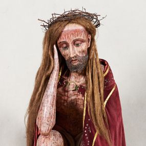Zdjęcie nr 1: Figura pełnoplastyczna przedstawiająca Chrystusa Frasobliwego. Chrystus ukazany w pozycji siedzącej na drewnianym postumencie, wspierający prawą ręką głowę, a lewą opierający na lewym udzie. Ciało o silnie podkreślonej klatce piersiowej z wąską talią, bardzo szczupłe. Twarz podłużna z długim i wąskim nosem, z głęboko osadzonymi oczami, wzrokiem skierowanym w dół, okolona gęstą, krótką brodą, zawiniętą na końcu w dwa małe pukle. Figura wyrzeźbiona bez włosów. Na głowę założona peruka z długich, naturalnych, jasnych włosów, a na nich korona z przeplecionych gałązek. Perizonium rzeźbione, białe, ciasno oplatające biodra, z jednym końcem rozwianym pod prawym udem. Polichromia w odsłoniętych partiach ciała naturalistyczna. Spod korony cierniowej oraz po całym ciele spływają bardzo liczne, drobne strużki krwi oraz zaznaczone poziomymi kreskami sińce. Na rzeźbę nałożony jest współczesny czerwony płaszcz z materiału, spięty pod szyją metalową klamrą, na brzegach obszyty złotą bordiurą. 


