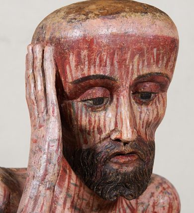 Zdjęcie nr 3: Figura pełnoplastyczna przedstawiająca Chrystusa Frasobliwego. Chrystus ukazany w pozycji siedzącej na drewnianym postumencie, wspierający prawą ręką głowę, a lewą opierający na lewym udzie. Ciało o silnie podkreślonej klatce piersiowej z wąską talią, bardzo szczupłe. Twarz podłużna z długim i wąskim nosem, z głęboko osadzonymi oczami, wzrokiem skierowanym w dół, okolona gęstą, krótką brodą, zawiniętą na końcu w dwa małe pukle. Figura wyrzeźbiona bez włosów. Na głowę założona peruka z długich, naturalnych, jasnych włosów, a na nich korona z przeplecionych gałązek. Perizonium rzeźbione, białe, ciasno oplatające biodra, z jednym końcem rozwianym pod prawym udem. Polichromia w odsłoniętych partiach ciała naturalistyczna. Spod korony cierniowej oraz po całym ciele spływają bardzo liczne, drobne strużki krwi oraz zaznaczone poziomymi kreskami sińce. Na rzeźbę nałożony jest współczesny czerwony płaszcz z materiału, spięty pod szyją metalową klamrą, na brzegach obszyty złotą bordiurą. 


