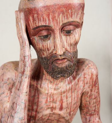 Zdjęcie nr 8: Figura pełnoplastyczna przedstawiająca Chrystusa Frasobliwego. Chrystus ukazany w pozycji siedzącej na drewnianym postumencie, wspierający prawą ręką głowę, a lewą opierający na lewym udzie. Ciało o silnie podkreślonej klatce piersiowej z wąską talią, bardzo szczupłe. Twarz podłużna z długim i wąskim nosem, z głęboko osadzonymi oczami, wzrokiem skierowanym w dół, okolona gęstą, krótką brodą, zawiniętą na końcu w dwa małe pukle. Figura wyrzeźbiona bez włosów. Na głowę założona peruka z długich, naturalnych, jasnych włosów, a na nich korona z przeplecionych gałązek. Perizonium rzeźbione, białe, ciasno oplatające biodra, z jednym końcem rozwianym pod prawym udem. Polichromia w odsłoniętych partiach ciała naturalistyczna. Spod korony cierniowej oraz po całym ciele spływają bardzo liczne, drobne strużki krwi oraz zaznaczone poziomymi kreskami sińce. Na rzeźbę nałożony jest współczesny czerwony płaszcz z materiału, spięty pod szyją metalową klamrą, na brzegach obszyty złotą bordiurą. 


