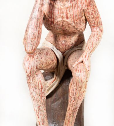 Zdjęcie nr 9: Figura pełnoplastyczna przedstawiająca Chrystusa Frasobliwego. Chrystus ukazany w pozycji siedzącej na drewnianym postumencie, wspierający prawą ręką głowę, a lewą opierający na lewym udzie. Ciało o silnie podkreślonej klatce piersiowej z wąską talią, bardzo szczupłe. Twarz podłużna z długim i wąskim nosem, z głęboko osadzonymi oczami, wzrokiem skierowanym w dół, okolona gęstą, krótką brodą, zawiniętą na końcu w dwa małe pukle. Figura wyrzeźbiona bez włosów. Na głowę założona peruka z długich, naturalnych, jasnych włosów, a na nich korona z przeplecionych gałązek. Perizonium rzeźbione, białe, ciasno oplatające biodra, z jednym końcem rozwianym pod prawym udem. Polichromia w odsłoniętych partiach ciała naturalistyczna. Spod korony cierniowej oraz po całym ciele spływają bardzo liczne, drobne strużki krwi oraz zaznaczone poziomymi kreskami sińce. Na rzeźbę nałożony jest współczesny czerwony płaszcz z materiału, spięty pod szyją metalową klamrą, na brzegach obszyty złotą bordiurą. 


