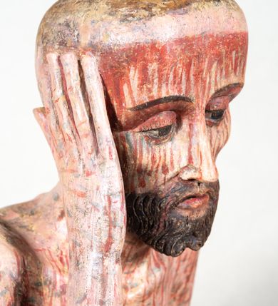 Zdjęcie nr 11: Figura pełnoplastyczna przedstawiająca Chrystusa Frasobliwego. Chrystus ukazany w pozycji siedzącej na drewnianym postumencie, wspierający prawą ręką głowę, a lewą opierający na lewym udzie. Ciało o silnie podkreślonej klatce piersiowej z wąską talią, bardzo szczupłe. Twarz podłużna z długim i wąskim nosem, z głęboko osadzonymi oczami, wzrokiem skierowanym w dół, okolona gęstą, krótką brodą, zawiniętą na końcu w dwa małe pukle. Figura wyrzeźbiona bez włosów. Na głowę założona peruka z długich, naturalnych, jasnych włosów, a na nich korona z przeplecionych gałązek. Perizonium rzeźbione, białe, ciasno oplatające biodra, z jednym końcem rozwianym pod prawym udem. Polichromia w odsłoniętych partiach ciała naturalistyczna. Spod korony cierniowej oraz po całym ciele spływają bardzo liczne, drobne strużki krwi oraz zaznaczone poziomymi kreskami sińce. Na rzeźbę nałożony jest współczesny czerwony płaszcz z materiału, spięty pod szyją metalową klamrą, na brzegach obszyty złotą bordiurą. 


