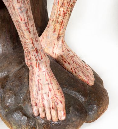 Zdjęcie nr 12: Figura pełnoplastyczna przedstawiająca Chrystusa Frasobliwego. Chrystus ukazany w pozycji siedzącej na drewnianym postumencie, wspierający prawą ręką głowę, a lewą opierający na lewym udzie. Ciało o silnie podkreślonej klatce piersiowej z wąską talią, bardzo szczupłe. Twarz podłużna z długim i wąskim nosem, z głęboko osadzonymi oczami, wzrokiem skierowanym w dół, okolona gęstą, krótką brodą, zawiniętą na końcu w dwa małe pukle. Figura wyrzeźbiona bez włosów. Na głowę założona peruka z długich, naturalnych, jasnych włosów, a na nich korona z przeplecionych gałązek. Perizonium rzeźbione, białe, ciasno oplatające biodra, z jednym końcem rozwianym pod prawym udem. Polichromia w odsłoniętych partiach ciała naturalistyczna. Spod korony cierniowej oraz po całym ciele spływają bardzo liczne, drobne strużki krwi oraz zaznaczone poziomymi kreskami sińce. Na rzeźbę nałożony jest współczesny czerwony płaszcz z materiału, spięty pod szyją metalową klamrą, na brzegach obszyty złotą bordiurą. 


