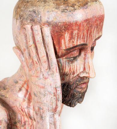 Zdjęcie nr 13: Figura pełnoplastyczna przedstawiająca Chrystusa Frasobliwego. Chrystus ukazany w pozycji siedzącej na drewnianym postumencie, wspierający prawą ręką głowę, a lewą opierający na lewym udzie. Ciało o silnie podkreślonej klatce piersiowej z wąską talią, bardzo szczupłe. Twarz podłużna z długim i wąskim nosem, z głęboko osadzonymi oczami, wzrokiem skierowanym w dół, okolona gęstą, krótką brodą, zawiniętą na końcu w dwa małe pukle. Figura wyrzeźbiona bez włosów. Na głowę założona peruka z długich, naturalnych, jasnych włosów, a na nich korona z przeplecionych gałązek. Perizonium rzeźbione, białe, ciasno oplatające biodra, z jednym końcem rozwianym pod prawym udem. Polichromia w odsłoniętych partiach ciała naturalistyczna. Spod korony cierniowej oraz po całym ciele spływają bardzo liczne, drobne strużki krwi oraz zaznaczone poziomymi kreskami sińce. Na rzeźbę nałożony jest współczesny czerwony płaszcz z materiału, spięty pod szyją metalową klamrą, na brzegach obszyty złotą bordiurą. 


