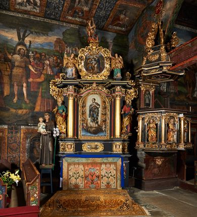 Zdjęcie nr 7: Obraz w kształcie owalu przedstawiający Świętą Rodzinę ukazaną na tle wnętrza. Po lewej stronie siedzący na krześle ustawionym na podeście św. Józef z Dzieciątkiem. Twarz świętego o rysach dojrzałego mężczyzny z długim nosem, okolona siwą brodą i włosami. Ubrany jest czerwone spodnie, szarą suknię, ugrowy płaszcz i sandały. Dzieciątko ukazane jest w pozycji siedzącej, zwrócone trzy czwarte w prawo z głową odchyloną w lewo i szeroko rozłożonymi rękami. Twarz o rysach dziecięcych, pulchnych i zarumienionych policzkach z podkreślonym podbródkiem; włosy kędzierzawe. Ubrane jest w białą sukienkę, przewiązaną czerwonym paskiem. Po prawej stronie obrazu Matka Boska wstępująca jedną nogą na podest, podtrzymująca obiema rękami Dzieciątko. Maria ukazana jest w pozycji stojącej, zwrócona trzy czwarte w prawo. Twarz pełna o jasnej karnacji, oczy przymknięte, nos długi i prosty, włosy upięte z tyłu głowy. Ubrana jest w długą, różową suknię, ciemnobłękitny płaszcz okrywający plecy i ramiona oraz sandały. Nad głowami wszystkich postaci koliste nimby. Powyżej grupy, pośród obłoków ukazane dwa nagie, uskrzydlone putta trzymające koronę, wieniec oraz lilię na tle obłoków. Postacie flankują dwa stojące we wnętrzu wazony z liliami. Kolorystyka przytłumiona, brunatna; szaty wydobyte światłocieniem.