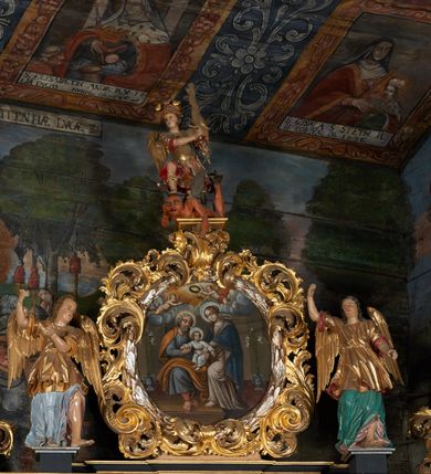 Zdjęcie nr 6: Obraz w kształcie owalu przedstawiający Świętą Rodzinę ukazaną na tle wnętrza. Po lewej stronie siedzący na krześle ustawionym na podeście św. Józef z Dzieciątkiem. Twarz świętego o rysach dojrzałego mężczyzny z długim nosem, okolona siwą brodą i włosami. Ubrany jest czerwone spodnie, szarą suknię, ugrowy płaszcz i sandały. Dzieciątko ukazane jest w pozycji siedzącej, zwrócone trzy czwarte w prawo z głową odchyloną w lewo i szeroko rozłożonymi rękami. Twarz o rysach dziecięcych, pulchnych i zarumienionych policzkach z podkreślonym podbródkiem; włosy kędzierzawe. Ubrane jest w białą sukienkę, przewiązaną czerwonym paskiem. Po prawej stronie obrazu Matka Boska wstępująca jedną nogą na podest, podtrzymująca obiema rękami Dzieciątko. Maria ukazana jest w pozycji stojącej, zwrócona trzy czwarte w prawo. Twarz pełna o jasnej karnacji, oczy przymknięte, nos długi i prosty, włosy upięte z tyłu głowy. Ubrana jest w długą, różową suknię, ciemnobłękitny płaszcz okrywający plecy i ramiona oraz sandały. Nad głowami wszystkich postaci koliste nimby. Powyżej grupy, pośród obłoków ukazane dwa nagie, uskrzydlone putta trzymające koronę, wieniec oraz lilię na tle obłoków. Postacie flankują dwa stojące we wnętrzu wazony z liliami. Kolorystyka przytłumiona, brunatna; szaty wydobyte światłocieniem.