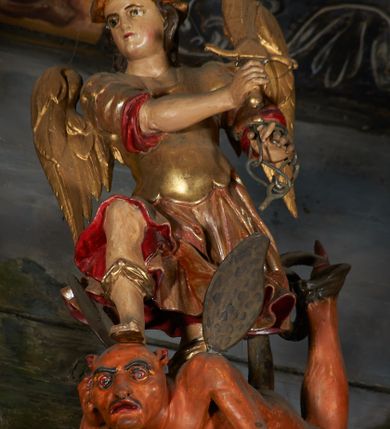 Zdjęcie nr 1: Rzeźba pełna przedstawiająca św. Michała Archanioła depczącego szatana. Archanioł zwrócony trzy czwarte w prawo, w pozycji stojącej z prawą nogą uniesioną, wspartą na głowie szatana. W prawej dłoni trzyma ognisty miecz, skierowany w górę, a w lewej wysuniętej do przodu fragment łańcucha od wagi. Twarz pełna, oczy szeroko otwarte, nos i usta niewielkie. Święty Michał ubrany jest w strój legionisty, złotą i krótką tunikę z czerwonym podbiciem, zbroję torsową oraz hełm z pióropuszem na głowie. Figura polichromowana jest naturalistycznie, szaty i atrybuty złocone. Pod jego stopami leży na brzuchu szatan z głową uniesioną, wspartą na prawej ręce, lewą rękę opuszcza w dół. Postać przedstawiona monstrualnie z kopytami i czarnym ogonem. Twarz owalna z dużymi i przekrwionymi oczami, czarnymi brwiami, wydatnym nosem i ustami rozchylonymi w lekkim grymasie, uszy małe i sterczące do góry. Szatan polichromowany w kolorze jasnobrązowym.