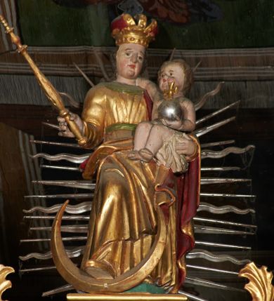 Zdjęcie nr 1: Rzeźba ścięta z tyłu, przedstawia Matkę Boską z Dzieciątkiem stojącą na niskim, zielonym, prostopadłościennym cokole o ściętych narożach. Maria ustawiona frontalnie w delikatnym kontrapoście z prawą stopą ustawioną na półksiężycu, berłem w prawej dłoni i Dzieciątkiem na lewym ręku. Twarz szeroka z pełnymi policzkami i wyraźnie zaznaczonym podbródkiem. Włosy długie, falowane, opadające na ramiona. Ubrana jest w długą, złoconą suknię przewiązaną zielonym paskiem, z zieloną chustą pod szyją oraz złocony płaszcz z czerwoną podszewką, odsłaniający prawe ramię i przerzucony prawą połą przez kolana na lewe przedramię. Na głowie ma założoną koronę zamkniętą. Dzieciątko zwrócone trzy czwarte w prawo, siedzące na białej pieluszce, prawą rączką obejmuje matkę za szyję, w lewej trzyma jabłko królewski. Twarz o rysach dziecięcych z małymi ustami i pełnymi policzkami. Włosy krótkie, brązowe, zasłaniające uszy. Rzeźby ustawione na tle mandorli o naprzemiennie prostych i falistych promieniach. 
