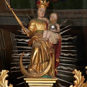 Zdjęcie nr 1: Rzeźba ścięta z tyłu, przedstawia Matkę Boską z Dzieciątkiem stojącą na niskim, zielonym, prostopadłościennym cokole o ściętych narożach. Maria ustawiona frontalnie w delikatnym kontrapoście z prawą stopą ustawioną na półksiężycu, berłem w prawej dłoni i Dzieciątkiem na lewym ręku. Twarz szeroka z pełnymi policzkami i wyraźnie zaznaczonym podbródkiem. Włosy długie, falowane, opadające na ramiona. Ubrana jest w długą, złoconą suknię przewiązaną zielonym paskiem, z zieloną chustą pod szyją oraz złocony płaszcz z czerwoną podszewką, odsłaniający prawe ramię i przerzucony prawą połą przez kolana na lewe przedramię. Na głowie ma założoną koronę zamkniętą. Dzieciątko zwrócone trzy czwarte w prawo, siedzące na białej pieluszce, prawą rączką obejmuje matkę za szyję, w lewej trzyma jabłko królewski. Twarz o rysach dziecięcych z małymi ustami i pełnymi policzkami. Włosy krótkie, brązowe, zasłaniające uszy. Rzeźby ustawione na tle mandorli o naprzemiennie prostych i falistych promieniach. 
