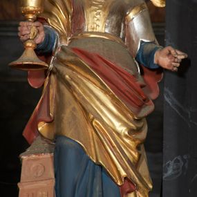 Zdjęcie nr 1: Rzeźba ścięta z tyłu, przedstawiająca św. Barbarę, ustawiona na niskim prostopadłościennym cokole o ściętych narożach. Święta ukazana frontalnie z głową skierowaną w lewą stronę. Lewą rękę unosi na wysokość pasa, w prawej trzyma kielich. Twarz okrągła o delikatnych rysach z wysokim czołem, drobnymi ustami i wyraźnie zaznaczonym podbródkiem. Włosy falowane z przedziałkiem na środku głowy, spływające  na ramiona. Święta ubrana jest w niebieską suknię spodnią, złotą suknię wierzchnią, przepasaną w talii; złoty płaszcz, podbity czerwienią, odsłaniający lewe ramię i zawinięty na prawym boku oraz brązowe buty. Przy prawej nodze świętej ustawiona jest miniaturowa trójkondygnacyjna wieża na rzucie kwadratu. Polichromia ciała naturalistyczna, detale złocone i srebrzone.