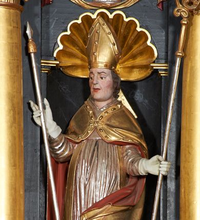 Zdjęcie nr 1: Rzeźba ścięta z tyłu, przedstawiająca św. Stanisława. Święty zwrócony jest  trzy czwarte w prawo, w delikatnym kontrapoście, z lewą nogą ugiętą w kolanie. W lewej ręce trzyma pastorał, a w prawej włócznię. Twarz szeroka o dużych półprzymkniętych oczach, dużym nosie i rozchylonych ustach. Włosy brązowe, średniej długości, zasłaniające uszy. Ubrany jest w szarą albę, srebrną komżę, złotą stułę oraz złotą kapę z czerwonym podbiciem, zapiętą na piersi, na brzegu zdobioną kaboszonami i rautami. Na głowie ma założoną infułę, na dłoniach białe rękawice, a na stopach czarne buty. Po prawej stronie świętego ustawiona jest niewielka rzeźba Piotrowina, ukazanego w pozycji klęczącej ze złożonymi w geście modlitwy dłońmi. Twarz szeroka z długim nosem, okolona wąsami i krótkimi włosami. Piotrowin ubrany jest w białą szatę z długimi rękawami. Polichromia rzeźb w odsłoniętych partiach ciała naturalistyczna, detale złocone i srebrzone.