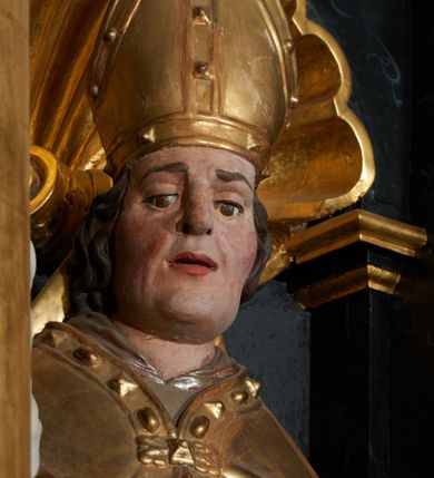 Zdjęcie nr 2: Rzeźba ścięta z tyłu, przedstawiająca św. Stanisława. Święty zwrócony jest  trzy czwarte w prawo, w delikatnym kontrapoście, z lewą nogą ugiętą w kolanie. W lewej ręce trzyma pastorał, a w prawej włócznię. Twarz szeroka o dużych półprzymkniętych oczach, dużym nosie i rozchylonych ustach. Włosy brązowe, średniej długości, zasłaniające uszy. Ubrany jest w szarą albę, srebrną komżę, złotą stułę oraz złotą kapę z czerwonym podbiciem, zapiętą na piersi, na brzegu zdobioną kaboszonami i rautami. Na głowie ma założoną infułę, na dłoniach białe rękawice, a na stopach czarne buty. Po prawej stronie świętego ustawiona jest niewielka rzeźba Piotrowina, ukazanego w pozycji klęczącej ze złożonymi w geście modlitwy dłońmi. Twarz szeroka z długim nosem, okolona wąsami i krótkimi włosami. Piotrowin ubrany jest w białą szatę z długimi rękawami. Polichromia rzeźb w odsłoniętych partiach ciała naturalistyczna, detale złocone i srebrzone.