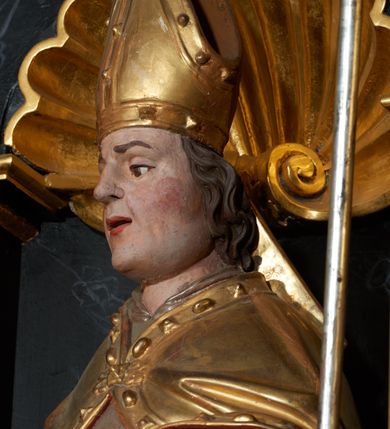 Zdjęcie nr 3: Rzeźba ścięta z tyłu, przedstawiająca św. Stanisława. Święty zwrócony jest  trzy czwarte w prawo, w delikatnym kontrapoście, z lewą nogą ugiętą w kolanie. W lewej ręce trzyma pastorał, a w prawej włócznię. Twarz szeroka o dużych półprzymkniętych oczach, dużym nosie i rozchylonych ustach. Włosy brązowe, średniej długości, zasłaniające uszy. Ubrany jest w szarą albę, srebrną komżę, złotą stułę oraz złotą kapę z czerwonym podbiciem, zapiętą na piersi, na brzegu zdobioną kaboszonami i rautami. Na głowie ma założoną infułę, na dłoniach białe rękawice, a na stopach czarne buty. Po prawej stronie świętego ustawiona jest niewielka rzeźba Piotrowina, ukazanego w pozycji klęczącej ze złożonymi w geście modlitwy dłońmi. Twarz szeroka z długim nosem, okolona wąsami i krótkimi włosami. Piotrowin ubrany jest w białą szatę z długimi rękawami. Polichromia rzeźb w odsłoniętych partiach ciała naturalistyczna, detale złocone i srebrzone.