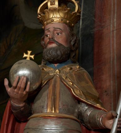 Zdjęcie nr 3: Rzeźba ścięta z tyłu, przedstawiająca św. Władysława. Święty ukazany frontalnie z jabłkiem królewskim w prawej dłoni i toporem w lewej. Twarz podłużna o głęboko osadzonych oczach i krótkim nosie, okolona brodą. Włosy półdługie, falowane. Święty ubrany jest w pełną, srebrną zbroję płytową, niebieską tunikę lamowaną złotem oraz złoty płaszcz spięty klamrą, podbity czerwienią, obficie drapowany w dolnej partii, w górnej zdobiony rautami i kaboszonami, na głowie ma założoną koronę zamkniętą. Polichromia naturalistyczna w partiach ciała, strój i atrybuty złocone i srebrzone.
