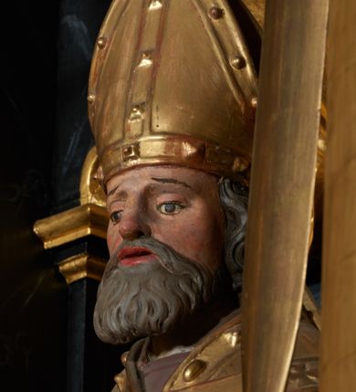 Zdjęcie nr 3: Rzeźba ścięta z tyłu, przedstawiająca św. Wojciecha. Święty ustawiony frontalnie, w lekkim kontrapoście z prawą nogą ugiętą w kolanie. W lewej ręce trzyma wiosło, a w prawej krzyż patriarchalny. Twarz o starczych rysach, na wpół przymkniętych oczach, rozchylonych ustach; okolona siwą brodą. Włosy siwe, średniej długości, zasłaniające uszy. Święty ubrany jest w fioletową albę, srebrną komżę, złotą kapę z czerwonym podbiciem, spiętą na piersi, złotą stułę, fioletowe rękawice, złotą infułę i brązowe buty. Kapa i infuła ozdobione są kaboszonami i rautami. Polichromia ciała naturalistyczna, szaty i atrybuty złocone i srebrzone.