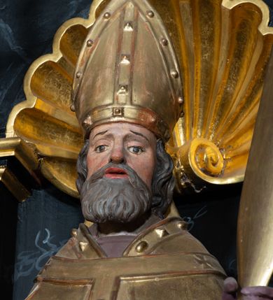 Zdjęcie nr 4: Rzeźba ścięta z tyłu, przedstawiająca św. Wojciecha. Święty ustawiony frontalnie, w lekkim kontrapoście z prawą nogą ugiętą w kolanie. W lewej ręce trzyma wiosło, a w prawej krzyż patriarchalny. Twarz o starczych rysach, na wpół przymkniętych oczach, rozchylonych ustach; okolona siwą brodą. Włosy siwe, średniej długości, zasłaniające uszy. Święty ubrany jest w fioletową albę, srebrną komżę, złotą kapę z czerwonym podbiciem, spiętą na piersi, złotą stułę, fioletowe rękawice, złotą infułę i brązowe buty. Kapa i infuła ozdobione są kaboszonami i rautami. Polichromia ciała naturalistyczna, szaty i atrybuty złocone i srebrzone.