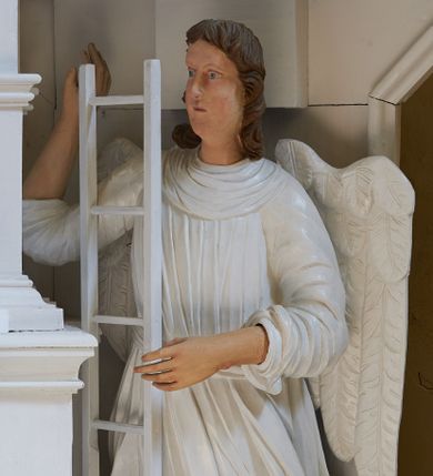 Zdjęcie nr 1: Rzeźba drążona, ustawiona na niewielkim postumencie. Figura zwrócona delikatnie w prawo, z prawą nogą ugiętą w kolanie, w obu rekach przytrzymuje wysoką drabinę. Twarz jest podłużna o pełnych policzkach oraz cofniętej brodzie; usta małe i delikatnie rozchylone, nos długi. Włosy są brązowe, falowane, sięgające do ramion. Anioł ubrany jest w długą, obficie marszczoną, białą szatę spodnią odsłaniającą prawą nogę oraz krótszą, białą szatę wierzchnią z podwiniętymi rękawami. Polichromia naturalistyczna w partiach ciała, szaty i atrybuty białe.