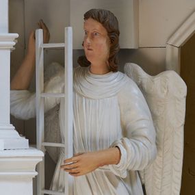 Zdjęcie nr 1: Rzeźba drążona, ustawiona na niewielkim postumencie. Figura zwrócona delikatnie w prawo, z prawą nogą ugiętą w kolanie, w obu rekach przytrzymuje wysoką drabinę. Twarz jest podłużna o pełnych policzkach oraz cofniętej brodzie; usta małe i delikatnie rozchylone, nos długi. Włosy są brązowe, falowane, sięgające do ramion. Anioł ubrany jest w długą, obficie marszczoną, białą szatę spodnią odsłaniającą prawą nogę oraz krótszą, białą szatę wierzchnią z podwiniętymi rękawami. Polichromia naturalistyczna w partiach ciała, szaty i atrybuty białe.