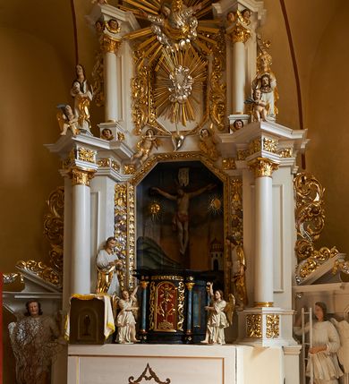 Zdjęcie nr 1: Rzeźba przedstawia św. Weronikę. Figura ustawiona na niewielkim, prostopadłościennym cokole. Święta ukazana w pozycji stojącej, frontalnie z rękami uniesionymi, w których trzyma veraikon. Twarz podłużna o pełnych policzkach i podbródku, z małymi i delikatnie otwartymi ustami, krótkim nosem i wyraźnie zaznaczonymi łukami brwiowymi, policzki silnie zarumienione. Święta ubrana jest w długą, białą suknię ze złoconą chustą u szyi oraz złocony płaszcz założony na głowę; na stopach ma założone białe buty. Veraikon w formie srebrzonej chusty z płaskorzeźbioną twarzą Chrystusa skierowaną na wprost. Twarz okolona krótką, ciemnobrązową brodą; włosy ułożone w dwa skręcone pukle, spływające do przodu. Spod zielonej korony cierniowej spływają strużki krwi. Polichromia naturalistyczna w partiach ciała, płaszcz złocony, chusta srebrzona.