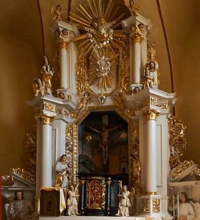 Zdjęcie nr 1: Nastawa ołtarzowa architektoniczna, jednoosiowa, dwukondygnacyjna, na cokole z bramkami. Ołtarz prostopadłościenny, drewniany; mensa drewniana. Antepedium w formie leżącego prostokąta, ujęte z trzech stron listwą o złoconym profilu, w centrum malowany złotem monogram maryjny. Tabernakulum w formie skrzynki o wklęsło-wypukłym wykroju, ustawione na profilowanym cokole, flankowane czterema kolumienkami o marmoryzowanych na niebiesko trzonach, podtrzymującymi niepełne belkowanie i wydatny gzyms. Ścianki boczne dekorowane prostokątnymi, marmoryzowanymi na niebiesko płycinami w złoconych ramach. Drzwiczki w kształcie stojącego prostokąta, zamkniętego łukiem nadwieszonym; w polu krucyfiks osadzony na wolutowej podstawie, od której odchodzą dwie gałązki akantu. Po bokach drzwiczek dwie wici roślinne. Tabernakulum flankują ustawione dwie figury aniołów.
Pierwsza kondygnacja retabulum ustawiona na cokole, ujęta parą filarów i stojących przed nimi, ustawionymi pod kątem kolumnami o kapitelach korynckich, dźwigających pełne belkowanie z wydatnym gzymsem. W polu środkowym rama w kształcie stojącego prostokąta zamkniętego trzema bokami z ośmioboku, dekorowana kampanulami, ujęta festonami z kampanulami, kwiatami i owocami. W polu obrazowym krucyfiks ukazany na malowanym tle Golgoty. Po bokach tabernakulum ustawione dwie figury: św. Jana Ewangelisty po lewej i św. Łukasza po prawej stronie.
Pośrodku belkowania zawieszone duże serce przebite sześcioma mieczami. Po jego bokach dwie rzeźby aniołków. Na gzymsie, na osi kolumny po prawej stronie ustawiona jest rzeźba św. Weroniki, a przed nią siedzący aniołek z trzema gwoździami w dłoni. Po lewej stronie św. Maria Magdalena, a przed nią aniołek z młotkiem w dłoni. 
Druga kondygnacja jest węższa, ujęta parą pilastrów i parą dostawionych pod kątem kolumn o kapitelach kompozytowych, dźwigających imposty. Cokoły kolumn i imposty ozdobione są z każdej strony uskrzydlonymi główkami aniołków. W polu rama w kształcie stojącego prostokąta o ściętych narożach, zdobiona ornamentem roślinnym, ujęta ornamentem z akantu i karbowanej wstęgi. Pośrodku promienista gloria z rzeźbioną gołębicą Ducha Świętego na tle obłoków. Całość zwieńczona drugą promienistą glorią z półpostacią Boga Ojca na tle obłoków. Po bokach na osi podpór siedzą dwie rzeźby aniołków. Obie kondygnacje ujęte są uszami utworzonymi z akantu i karbowanej wstęgi.
Bramki z przejściami w formie stojącego prostokąta o ściętych górnych narożach, zamknięte wydatnym gzymsem dźwiganym przez fragmenty pilastrów, na ich osi przerwany trójkątny przyczółek, zdobiony akantem. Przejścia flankowane figurami aniołów z arma Christi, nad bramkami figury proroków.
Struktura polichromowana na biało, ornamentyka i detale architektoniczne złocone i srebrzone.

