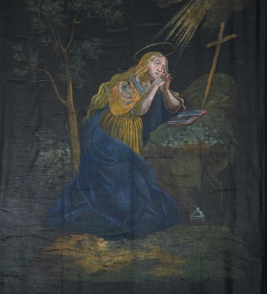 Zdjęcie nr 1: Opona wielkopostna z przedstawieniem św. Marii Magdaleny. W centrum kompozycji ukazana święta klęcząca przed skałą, zwrócona trzy czwarte w lewo, z dłońmi splecionymi w geście modlitwy. Twarz pełna o młodzieńczych rysach i dużym nosie. Włosy jasne, długie, opadają na plecy i ramiona. Ubrana jest w żółtą, obficie drapowana suknię, przewiązana w talii oraz przerzucony przez lewe ramię i otaczający postać niebieski płaszcz, który opada obfitą draperią na ziemię. Święta ukazana jest na tle krajobrazu, u jej stóp postawiona jest puszka na wonności. Na skale leży otwarta księga, a obok stoi wysoki krzyż. Za świętą, po lewej stronie obrazu jest wysokie drzewo. W prawym górnym narożu obrazu znajdują się obłoki, z których wychodzą wiązki promieni kierowane ku świętej, a nad jej głową znajduje się złoty i okrągły nimb. Kolorystyka przygaszona, postać rozświetlona, o intensywnych plamach barwnych.