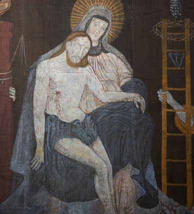 Zdjęcie nr 5: Tkanina wielkopostna przedstawiająca Piętę ukazaną na zszytych fragmentach płótna (25 brytów). W centrum kompozycji stoi krzyż, pod którym siedzi Matka Boska trzymająca na kolanach martwe ciało Chrystusa. Maria zwrócona jest trzy czwarte w lewo,  lewą ręką podtrzymuje rękę syna, prawą obejmuje go w talii, głowę ma przechyloną w lewo. Twarz podłużna o wyraźnych, konturowych rysach, oczy niemal zamknięte, długi nos, wąskie usta. Ubrana jest w jasną suknię o fałdach podkreślonych wyraźnymi konturami, niebieski płaszcz, okrywający nogi, podwikę oraz szary welon na głowie. Wokół głowy ma kolisty, złoty i promienisty nimb. Ciało Chrystusa spoczywa na kolanach Marii. Twarz owalna o konturowych rysach z zamkniętymi oczami, okolona jasnym zarostem, włosy długie z przedziałkiem na środku głowy, opadające na plecy. Ciało blade, ociekające krwią, niemodelowane, podkreślone konturem. Przez biodra przewiązane granatowe perizonium. O ramiona krzyża oparte ukośnie: po lewej włócznia, po prawej gąbka z octem na długim drzewcu. U góry krzyża zawieszony biały titulus w formie banderoli z tytułem winy w trzech językach „INRI”. Po bokach kompozycji u dołu, dwa anioły ukazane w pozycji klęczącej, zwrócone trzy czwarte ku środkowi obrazu. Anioł po prawej stronie trzyma w prawej dłoni drabinę, na której koniec założona jest korona cierniowa, nieco niżej ukazano trzy gwoździe, a wyżej rękawicę żołnierza. Anioł po lewej obejmuje kolumnę biczowania, na której stoi piejący kogut. W górnej części kolumny przywiązane są bicze. Ponad aniołami unoszą się na obłokach dwie kolejne postacie aniołów. Anioły ubrane są w różowe suknie i niebieskie tuniki z żółtą podszewką, przewiązane w pasie złotą taśmą. W górnych narożach obrazu symbole lunarne o ludzkich twarzach: po lewej księżyc, po prawej słońce. Na dole obrazu, pośrodku malowana na czerwono sygnatura „AB”. Po lewej stronie obrazu na wysokości ręki anioła unoszącego się na obłoku fragment daty „16”. Po bokach krzyża kilkuwersowe napisy, słabo czytelne. Według literatury nad głową Matki Boskiej znajduje się napis: „Filia populi ei acingere cilicio recospergere cinere: iuctu Unigenliafe tibi planctu emaru”.  Obok słońca i księżyca kolejny napis: „Ecce videntes clamabunt foris angeli pacis amare flebunt”, a także „amare flebunt angeli pacis”. Kolorystyka blada, w odcieniach zimnych, formy wydobyte konturem.