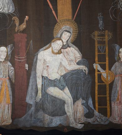 Zdjęcie nr 9: Tkanina wielkopostna przedstawiająca Piętę ukazaną na zszytych fragmentach płótna (25 brytów). W centrum kompozycji stoi krzyż, pod którym siedzi Matka Boska trzymająca na kolanach martwe ciało Chrystusa. Maria zwrócona jest trzy czwarte w lewo,  lewą ręką podtrzymuje rękę syna, prawą obejmuje go w talii, głowę ma przechyloną w lewo. Twarz podłużna o wyraźnych, konturowych rysach, oczy niemal zamknięte, długi nos, wąskie usta. Ubrana jest w jasną suknię o fałdach podkreślonych wyraźnymi konturami, niebieski płaszcz, okrywający nogi, podwikę oraz szary welon na głowie. Wokół głowy ma kolisty, złoty i promienisty nimb. Ciało Chrystusa spoczywa na kolanach Marii. Twarz owalna o konturowych rysach z zamkniętymi oczami, okolona jasnym zarostem, włosy długie z przedziałkiem na środku głowy, opadające na plecy. Ciało blade, ociekające krwią, niemodelowane, podkreślone konturem. Przez biodra przewiązane granatowe perizonium. O ramiona krzyża oparte ukośnie: po lewej włócznia, po prawej gąbka z octem na długim drzewcu. U góry krzyża zawieszony biały titulus w formie banderoli z tytułem winy w trzech językach „INRI”. Po bokach kompozycji u dołu, dwa anioły ukazane w pozycji klęczącej, zwrócone trzy czwarte ku środkowi obrazu. Anioł po prawej stronie trzyma w prawej dłoni drabinę, na której koniec założona jest korona cierniowa, nieco niżej ukazano trzy gwoździe, a wyżej rękawicę żołnierza. Anioł po lewej obejmuje kolumnę biczowania, na której stoi piejący kogut. W górnej części kolumny przywiązane są bicze. Ponad aniołami unoszą się na obłokach dwie kolejne postacie aniołów. Anioły ubrane są w różowe suknie i niebieskie tuniki z żółtą podszewką, przewiązane w pasie złotą taśmą. W górnych narożach obrazu symbole lunarne o ludzkich twarzach: po lewej księżyc, po prawej słońce. Na dole obrazu, pośrodku malowana na czerwono sygnatura „AB”. Po lewej stronie obrazu na wysokości ręki anioła unoszącego się na obłoku fragment daty „16”. Po bokach krzyża kilkuwersowe napisy, słabo czytelne. Według literatury nad głową Matki Boskiej znajduje się napis: „Filia populi ei acingere cilicio recospergere cinere: iuctu Unigenliafe tibi planctu emaru”.  Obok słońca i księżyca kolejny napis: „Ecce videntes clamabunt foris angeli pacis amare flebunt”, a także „amare flebunt angeli pacis”. Kolorystyka blada, w odcieniach zimnych, formy wydobyte konturem.