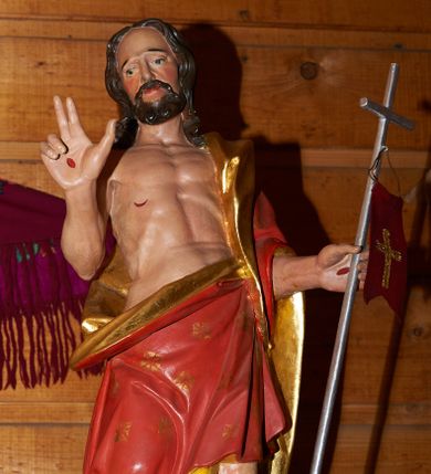 Zdjęcie nr 1: Rzeźba Chrystusa Zmartwychwstałego, pełna, ustawiona na kolistymi zielonym cokole. Chrystus ukazany  frontalnie, w kontrapoście, delikatnie przechylony w prawą stronę, w lewej ręce trzyma chorągiew zakończoną krzyżem, a prawą błogosławi. Twarz pociągła z wysokim czołem, oczy skierowane w dół, długi i wąski nos, broda średniej długości, ciemnobrązowa. Włosy długie, brązowe, z przedziałkiem na środku głowy, opadają na ramiona w postaci loków. Chrystus ubrany jest w czerwony płaszcz w złote gwiazdy, ze złotym podbiciem, przewieszony przez lewe ramię i zawiązany na lewym boku i odsłaniający nagi tors. Ciało o podkreślonej muskulaturze z zaznaczonymi śladami męki. Polichromia naturalistyczna, detal płaszcza złocony.