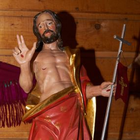 Zdjęcie nr 1: Rzeźba Chrystusa Zmartwychwstałego, pełna, ustawiona na kolistymi zielonym cokole. Chrystus ukazany  frontalnie, w kontrapoście, delikatnie przechylony w prawą stronę, w lewej ręce trzyma chorągiew zakończoną krzyżem, a prawą błogosławi. Twarz pociągła z wysokim czołem, oczy skierowane w dół, długi i wąski nos, broda średniej długości, ciemnobrązowa. Włosy długie, brązowe, z przedziałkiem na środku głowy, opadają na ramiona w postaci loków. Chrystus ubrany jest w czerwony płaszcz w złote gwiazdy, ze złotym podbiciem, przewieszony przez lewe ramię i zawiązany na lewym boku i odsłaniający nagi tors. Ciało o podkreślonej muskulaturze z zaznaczonymi śladami męki. Polichromia naturalistyczna, detal płaszcza złocony.