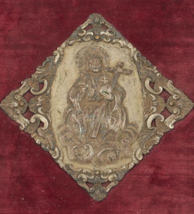 Zdjęcie nr 1: Okucia w kształcie częściowo ażurowych plakiet rozmieszczone na oprawie mszału wydanego w Wenecji w 1768 roku, na tle desek obciągniętych ciemnoczerwonym aksamitem. W narożach górnej i dolnej okładki trójkątne plakiety z motywami uskrzydlonych główek aniołów i ornamentem małżowinowym; przy krawędziach gładkie listwy łączące plakiety. Przy prawej krawędzi górnej okładziny i przy lewej krawędzi okładziny dolnej szyldziki zapięcia w formie bukietów kwiatów wyrastających z masek. Pośrodku okładzin rombowe plakiety z przedstawieniami – na górnej okładzinie Chrystus Salwator siedzący na obłokach, na dolnej Arma Christi; w narożach grzebienie ornamentu rocaille’owego (?) oraz motywy muszli i wolut. 