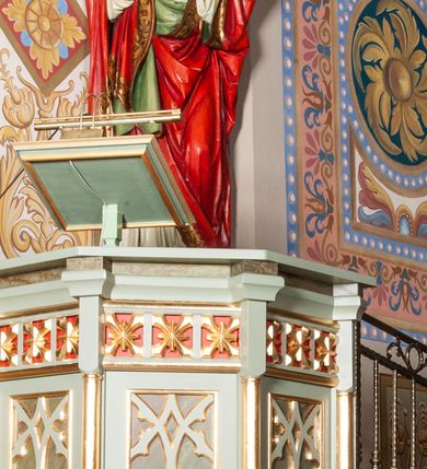 Zdjęcie nr 1: Ambona stojąca, ustawiona w prezbiterium. Stopa na planie ośmioboku z uskokiem; tuleja na podwójnym cokole, ośmioboczna, przechodząca w podstawę dla kosza; dekorowana w narożach maswerkiem. Kosz na planie sześciu boków z ośmioboku. Ścianki wydzielone wąskimi kolumienkami na wysokich cokołach podtrzymujących wysokie imposty i parapet kosza. W polu ścianek bocznych prostokątne płyciny z maswerkiem, powyżej fryz z kwadratowymi płycinami ze stylizowanymi rozetkami. Kosz u spodu dekorowany motywem geometrycznym i krenelażem. Na parapecie ustawiony na nóżce kwadratowy pulpit w profilowanej ramie. Z boku dwustopniowe schodki z metalową, ażurową balustradą, dekorowaną motywem roślinnym. Polichromia w kolorze jasnozielonym, z marmoryzowanymi i czerwonymi elementami, detale złocone.
