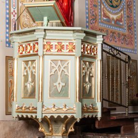 Zdjęcie nr 1: Ambona stojąca, ustawiona w prezbiterium. Stopa na planie ośmioboku z uskokiem; tuleja na podwójnym cokole, ośmioboczna, przechodząca w podstawę dla kosza; dekorowana w narożach maswerkiem. Kosz na planie sześciu boków z ośmioboku. Ścianki wydzielone wąskimi kolumienkami na wysokich cokołach podtrzymujących wysokie imposty i parapet kosza. W polu ścianek bocznych prostokątne płyciny z maswerkiem, powyżej fryz z kwadratowymi płycinami ze stylizowanymi rozetkami. Kosz u spodu dekorowany motywem geometrycznym i krenelażem. Na parapecie ustawiony na nóżce kwadratowy pulpit w profilowanej ramie. Z boku dwustopniowe schodki z metalową, ażurową balustradą, dekorowaną motywem roślinnym. Polichromia w kolorze jasnozielonym, z marmoryzowanymi i czerwonymi elementami, detale złocone.
