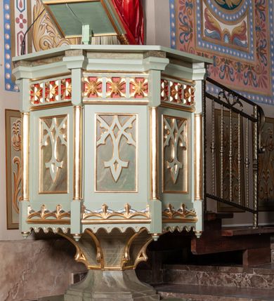 Zdjęcie nr 2: Ambona stojąca, ustawiona w prezbiterium. Stopa na planie ośmioboku z uskokiem; tuleja na podwójnym cokole, ośmioboczna, przechodząca w podstawę dla kosza; dekorowana w narożach maswerkiem. Kosz na planie sześciu boków z ośmioboku. Ścianki wydzielone wąskimi kolumienkami na wysokich cokołach podtrzymujących wysokie imposty i parapet kosza. W polu ścianek bocznych prostokątne płyciny z maswerkiem, powyżej fryz z kwadratowymi płycinami ze stylizowanymi rozetkami. Kosz u spodu dekorowany motywem geometrycznym i krenelażem. Na parapecie ustawiony na nóżce kwadratowy pulpit w profilowanej ramie. Z boku dwustopniowe schodki z metalową, ażurową balustradą, dekorowaną motywem roślinnym. Polichromia w kolorze jasnozielonym, z marmoryzowanymi i czerwonymi elementami, detale złocone.
