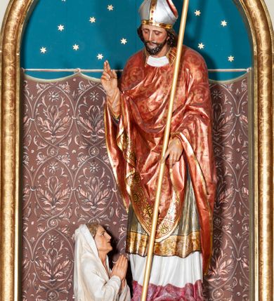 Zdjęcie nr 2: Rzeźba pełnoplastyczna przedstawiająca św. Stanisława Biskupa. Święty zwrócony jest delikatnie w prawo, pochylony delikatnie w dół w kierunku półpostaci Piotrowina. Prawą rękę wznosi ku górze w geście błogosławieństwa, w lewej opuszczonej w dół trzyma pastorał. Twarz trójkątna okolona ciemnym zarostem i długimi włosami spływającymi na plecy. Święty ubrany jest w białą albę z czerwonym pasem u dołu, wzorzysty ornat zdobiony szeroką bordiurą oraz czerwony ornat na planie koła dekorowany wicią roślinną; na głowie ma założoną srebrną infułę. U stóp biskupa, po lewej stronie ukazane jest popiersie Piotrowina przedstawione z prawego profilu, w białej szacie, z rękoma złożonymi w geście modlitewnym. Twarz szeroka z silnie wysuniętym podbródkiem z jasnym włosami schowanymi pod szatą, założoną na głowę. Polichromia w odsłoniętych partiach ciała naturalistyczna, detale złocone i srebrzone.