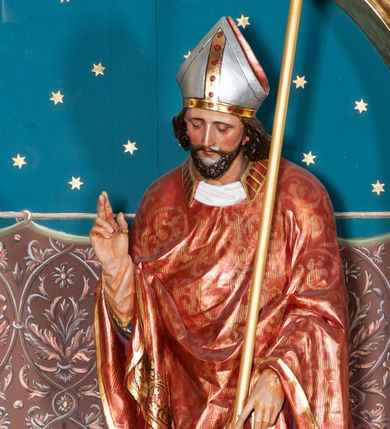 Zdjęcie nr 1: Rzeźba pełnoplastyczna przedstawiająca św. Stanisława Biskupa. Święty zwrócony jest delikatnie w prawo, pochylony delikatnie w dół w kierunku półpostaci Piotrowina. Prawą rękę wznosi ku górze w geście błogosławieństwa, w lewej opuszczonej w dół trzyma pastorał. Twarz trójkątna okolona ciemnym zarostem i długimi włosami spływającymi na plecy. Święty ubrany jest w białą albę z czerwonym pasem u dołu, wzorzysty ornat zdobiony szeroką bordiurą oraz czerwony ornat na planie koła dekorowany wicią roślinną; na głowie ma założoną srebrną infułę. U stóp biskupa, po lewej stronie ukazane jest popiersie Piotrowina przedstawione z prawego profilu, w białej szacie, z rękoma złożonymi w geście modlitewnym. Twarz szeroka z silnie wysuniętym podbródkiem z jasnym włosami schowanymi pod szatą, założoną na głowę. Polichromia w odsłoniętych partiach ciała naturalistyczna, detale złocone i srebrzone.