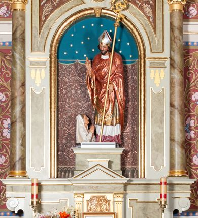 Zdjęcie nr 3: Rzeźba pełnoplastyczna przedstawiająca św. Stanisława Biskupa. Święty zwrócony jest delikatnie w prawo, pochylony delikatnie w dół w kierunku półpostaci Piotrowina. Prawą rękę wznosi ku górze w geście błogosławieństwa, w lewej opuszczonej w dół trzyma pastorał. Twarz trójkątna okolona ciemnym zarostem i długimi włosami spływającymi na plecy. Święty ubrany jest w białą albę z czerwonym pasem u dołu, wzorzysty ornat zdobiony szeroką bordiurą oraz czerwony ornat na planie koła dekorowany wicią roślinną; na głowie ma założoną srebrną infułę. U stóp biskupa, po lewej stronie ukazane jest popiersie Piotrowina przedstawione z prawego profilu, w białej szacie, z rękoma złożonymi w geście modlitewnym. Twarz szeroka z silnie wysuniętym podbródkiem z jasnym włosami schowanymi pod szatą, założoną na głowę. Polichromia w odsłoniętych partiach ciała naturalistyczna, detale złocone i srebrzone.