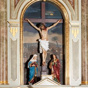 Zdjęcie nr 1: Ołtarz przyścienny, architektoniczny, jednoosiowy, jednokondygnacyjny. Mensa skrzyniowa, prostopadłościenna, z kamiennym portatylem, ustawiona na niskiej podstawie, antepedium ujęte płaskimi listwami łączącymi się z owalnym polem, pośrodku którego jest aplikowany, pozłacany krzyż. Predella wysoka, ujęta po bokach konsolami w formie uproszczonych wolut z nieznacznie wysuniętym architektonicznym tabernakulum, ujętym kanelowanymi pilastrami, podtrzymującymi belkowanie i trójkątny przyczółek. Pomiędzy pilastrami drzwiczki prostokątne, w których płycina w formie prostokąta o wklęsłych ćwierćkoliście narożach z malowanym ornamentem akantowym i kielichem z hostią pośrodku. Po bokach tabernakulum dwie marmoryzowane płyciny w kształcie kwadratu z uszakami. Nastawa ołtarzowa flankowana dwiema kolumnami jońskimi wspierającymi belkowanie z silnie wysuniętym gzymsem. Pole środkowe w formie głębokiej, prostokątnej, zamkniętej półkoliście wnęki o dekoracyjnej ramie, wewnątrz której umieszczona jest rzeźbiona grupa Ukrzyżowania na tle malowanego widoku Jerozolimy. Pola pomiędzy wnęką a kolumnami wypełnione są stylizowaną, profilowaną arkadą z archiwoltą wspartą na toskańskich kapitelach, pod którymi podwieszone są łezki. W górnej części arkady, po bokach znajdują się dwie płyciny wypełnione malowaną, białą  dekoracją roślinną na brązowym tle, a pomiędzy nimi jest w owalu rzeźbiona i złocona rozeta. Całość struktury ołtarza polichromowana i marmoryzowana jest w odcieniach beżu i szarości.