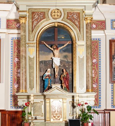 Zdjęcie nr 2: Ołtarz przyścienny, architektoniczny, jednoosiowy, jednokondygnacyjny. Mensa skrzyniowa, prostopadłościenna, z kamiennym portatylem, ustawiona na niskiej podstawie, antepedium ujęte płaskimi listwami łączącymi się z owalnym polem, pośrodku którego jest aplikowany, pozłacany krzyż. Predella wysoka, ujęta po bokach konsolami w formie uproszczonych wolut z nieznacznie wysuniętym architektonicznym tabernakulum, ujętym kanelowanymi pilastrami, podtrzymującymi belkowanie i trójkątny przyczółek. Pomiędzy pilastrami drzwiczki prostokątne, w których płycina w formie prostokąta o wklęsłych ćwierćkoliście narożach z malowanym ornamentem akantowym i kielichem z hostią pośrodku. Po bokach tabernakulum dwie marmoryzowane płyciny w kształcie kwadratu z uszakami. Nastawa ołtarzowa flankowana dwiema kolumnami jońskimi wspierającymi belkowanie z silnie wysuniętym gzymsem. Pole środkowe w formie głębokiej, prostokątnej, zamkniętej półkoliście wnęki o dekoracyjnej ramie, wewnątrz której umieszczona jest rzeźbiona grupa Ukrzyżowania na tle malowanego widoku Jerozolimy. Pola pomiędzy wnęką a kolumnami wypełnione są stylizowaną, profilowaną arkadą z archiwoltą wspartą na toskańskich kapitelach, pod którymi podwieszone są łezki. W górnej części arkady, po bokach znajdują się dwie płyciny wypełnione malowaną, białą  dekoracją roślinną na brązowym tle, a pomiędzy nimi jest w owalu rzeźbiona i złocona rozeta. Całość struktury ołtarza polichromowana i marmoryzowana jest w odcieniach beżu i szarości.