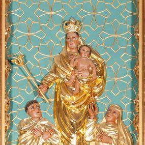 Zdjęcie nr 1: Kompozycja w kształcie stojącego prostokąta, w centrum płaskorzeźba z przedstawieniem Matki Boskiej stojącej na obłoku, na którym leżą jabłko królewskie i berło. Maria na lewym ręku trzyma Dzieciątko, a w prawej wyciągniętej przed siebie dzierży berło. Ubrana jest w suknię oraz w ściśle okalający jej ciało płaszcz; na głowie ma koronę zamkniętą. Twarz o delikatnych rysach, owalna. Dzieciątko ukazane jest w skręcie sylwetki, torsem zwrócone frontalnie; prawą rączka błogosławi, lewą podtrzymuje różaniec. U dołu klęczący św. Dominik oraz św. Katarzyna Sieneńska.  Po lewej św. Dominik zwrócony trzy czwarte w lewo, z głową skierowaną w górę oraz dłońmi skrzyżowanymi na piersi. Ubrany jest w habit i płaszcz dominikański , na głowie ma tonsurę. Po prawej stronie św. Katarzyna Sieneńska o analogicznym układzie ciała, prawą rękę wyciąga ku Dzieciątku, sięgając po różaniec, lewą wyciąga przed siebie. Ubrana jest w habit i płaszcz dominikański. Całość ukazana na zielonym tle, pokrytym rzeźbioną, złoconą, nieregularną kratką z geometrycznym motywem krzyży. Polichromia w partiach ciała naturalistyczna, szaty i atrybuty złocone, obłok srebrzony.