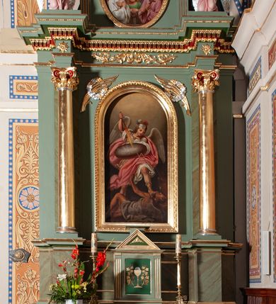 Zdjęcie nr 2: Obraz w kształcie stojącego prostokąta, zamkniętego łukiem półkolistym. W centrum kompozycji znajduje się św. Michał Archanioł w dynamicznej pozie, strącający szatana do piekła. Archanioł ukazany w postawie stojącej, z prawą nogą cofniętą do tyłu, lewą depczący szatana, z podniesioną prawą ręką do góry, w której trzyma płomienisty miecz skierowany ostrzem w dół, a w lewej owalną tarczę z napisem „KTÓŻ JAK BÓG?”. Od miecza biegnie zygzakiem piorun w stronę szatana. Twarz archanioła trójkątna, z długim i prostym nosem, wzrokiem skierowanym w dół. Włosy średniej długości, kręcone, rozwiane do tyłu. Archanioł ubrany jest w zbroję legionisty rzymskiego: ma białą tunikę obszytą złotą lamówką na brzegu, srebrny pancerz, hełm z trójkolorowym pióropuszem, rzymskie sandały oraz czerwony płaszcz rozwiany na wietrze; natomiast na plecach ma białe skrzydła. Na dole kompozycji ukazany jest szatan w pozycji leżącej z głową z prawej strony obrazu, lewą rękę wznoszący ku górze. Twarz owalna z rogami na czole, karnacja ciemna. Ubrany jest w białą szatę, odsłaniającą nagie ramiona i nogi; na plecach ma szare skrzydła nietoperza. Tło jednolite, szare, rozjaśnione w górnej części obrazu. Rama drewniana, złocona, od wewnątrz profilowana, od zewnątrz zdobiona astragalem.
